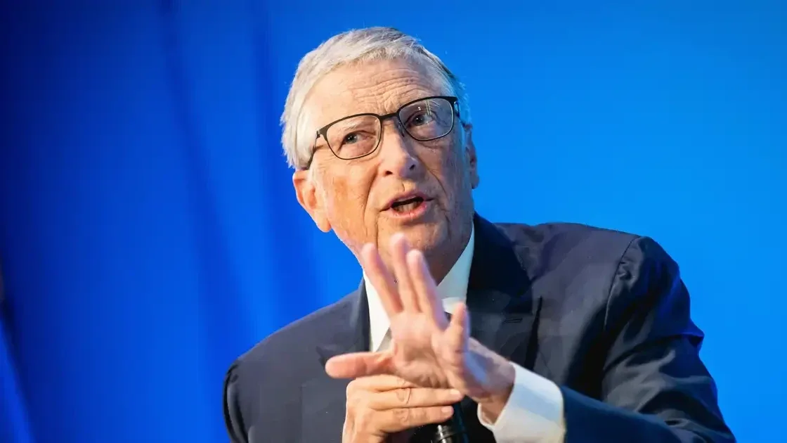 Bill Gates gesturing with an open hand while speaking, against a blue backdrop